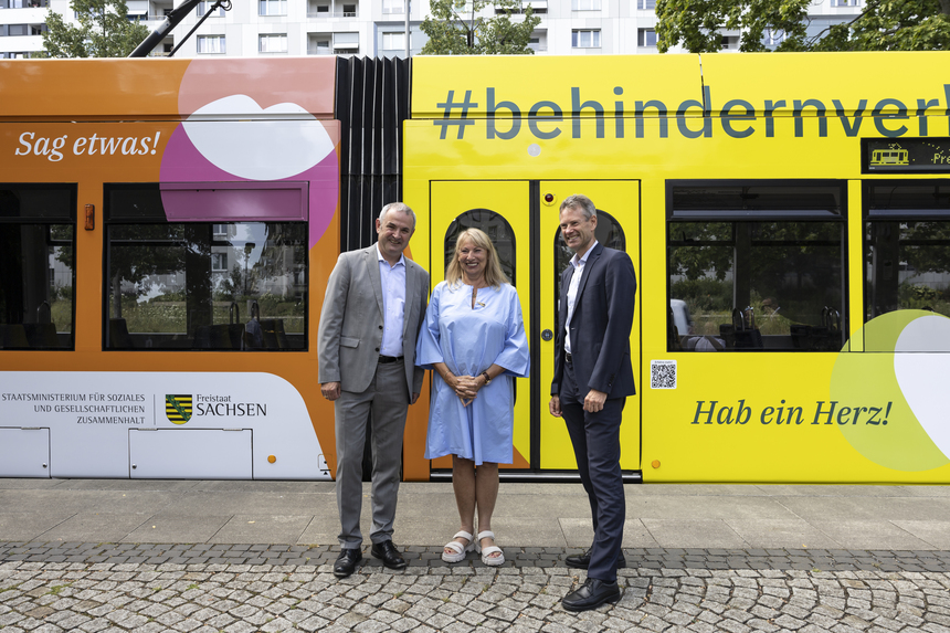 Der Landesbeauftragte Michael Welsch steht zusammen mit Frau Staatsministerin Petra Köpping und dem DVB-Vorstand Andreas Hemmersbach vor einer Straßenbahn, die mit den Motiven der Kampagne "Behindern.Verhindern" beklebt ist.