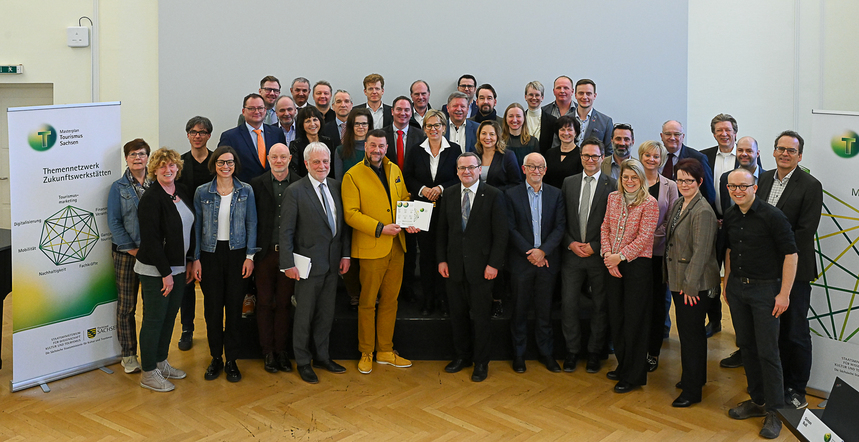 Die Teilnehmer der ersten Sitzung des Beirats zum Masterplan Tourismus Sachsen stehen als Gruppe in einem großen Raum vor einer weißen Wand und blicken in die Kamera.