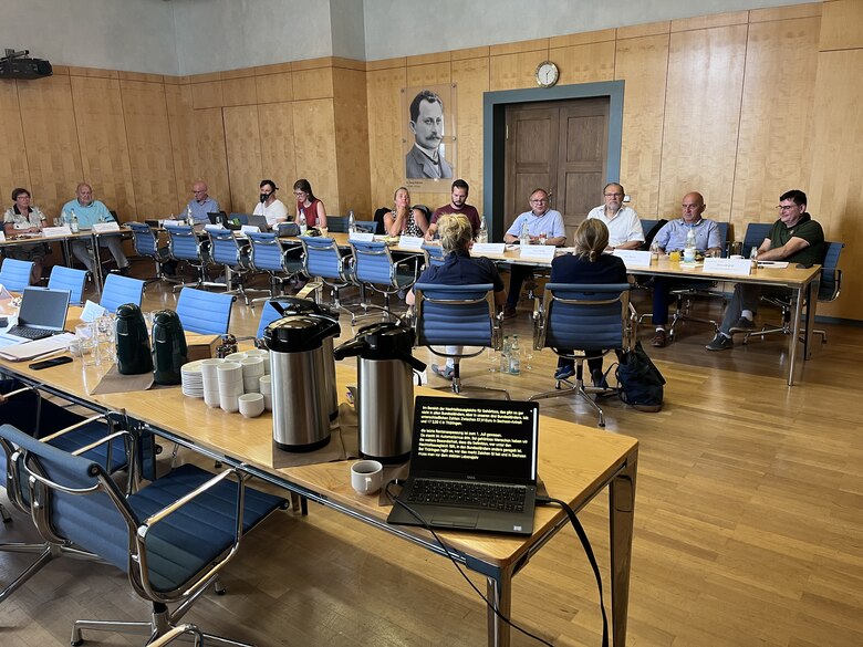 Teilnehmer des Mitteldeutschen Treffens der Landesbehindertenbeauftragten von Sachsen-Anhalt, Thüringen und Sachsen 2023 in der Sächsischen Staatskanzlei sitzen in einem Sitzungssaal an langen Tischen und sprechen miteinander. 