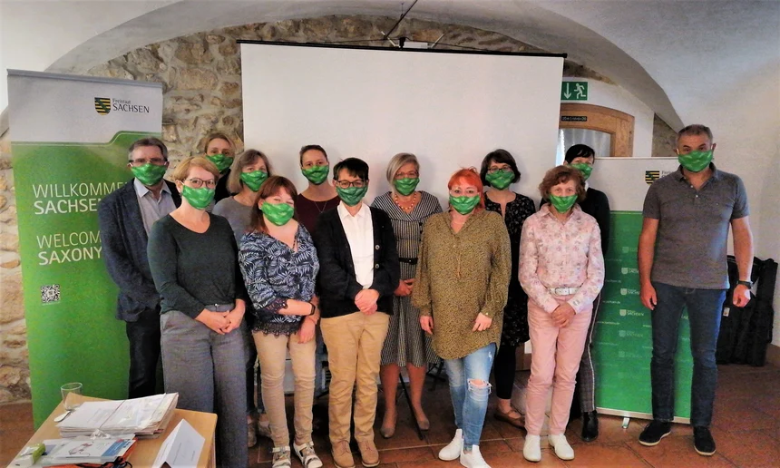 Die Beauftragten für Menschen mit Behinderungen der Landkreise und kreisfreien Städte stehen als Gruppe zusammen vor einer Wand. Alle tragen einen grünen Mund-Nase-Schutz. 