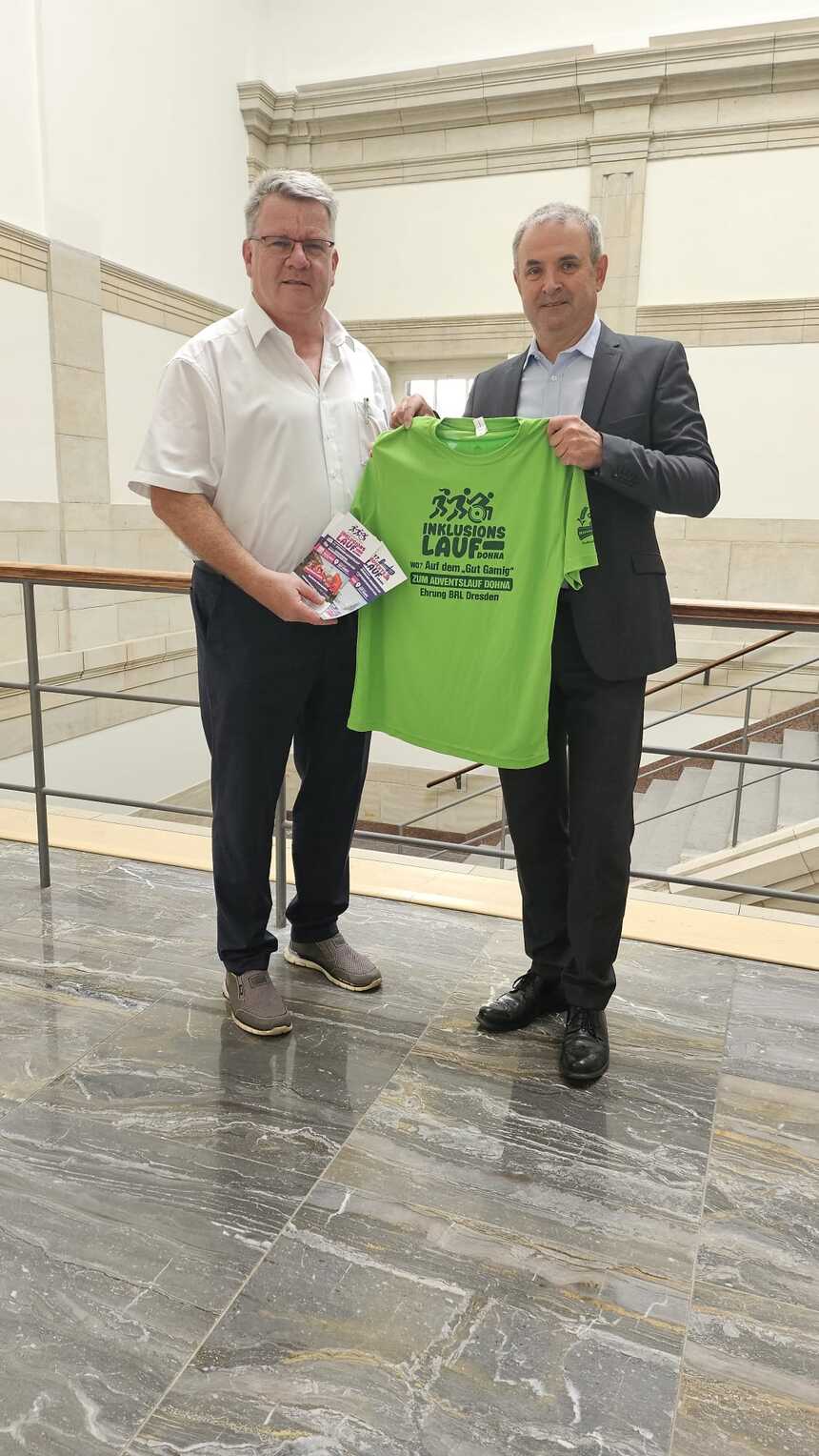 Der Landesbeauftragte Welsch erhält vom Dohnaer Bürgermeister Müller das offizielle Laufshirt für den 3. Sächsischen Inklusionslauf 2025 überreicht.