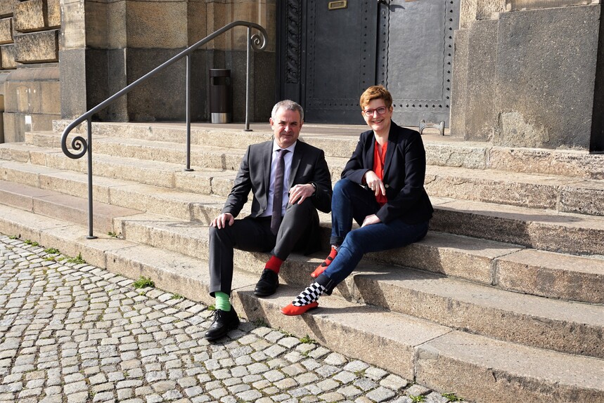 Die beiden Beauftragten der Sächsischen Staatsregierung, Herr Welsch und Frau Rüthrich, sitzen vor der Sächischen Staatskanzlei mit bunten Socken auf einer Treppe. 