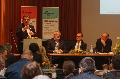 Foto: Der Beauftragte Stephan Pöhler steht während der Podiumsdiskussion am Rednerpult und spricht zu den Teilnehmern der Offenen Tagung.